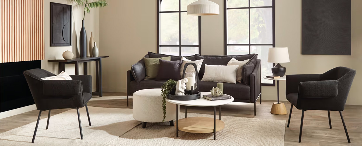 Room with beige walls, two windows, a dark brown sofa and chairs. The floor is light brown wood with a cream rug. In the center is a cream and wood table with various decor on top.