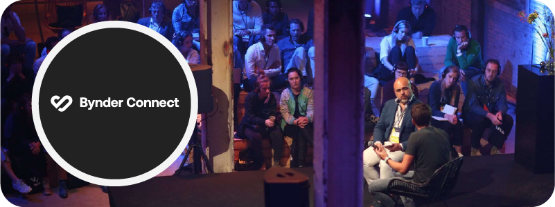 Image of people watching a presentation at Bynder Connect. The lighting is dim. The image has a purple hue. The presenters are highlighted with a spotlight as others sit around them. Everyone is in chairs. On the left side is a dark circle with Bynder Connect inside.